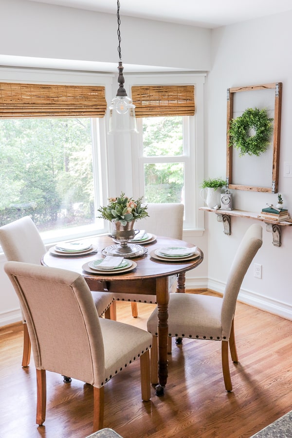 eat in kitchen with vintage round wooden table, cream parsons chairs next to a bay window with woven shades and farmhouse decor and table setting