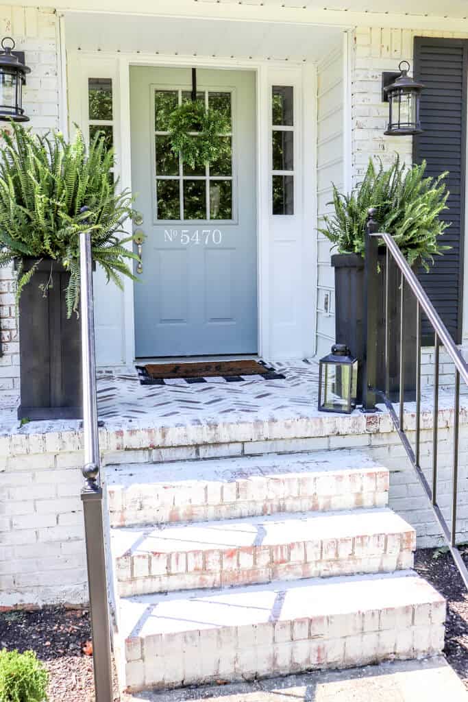 Limewashed brick home with a blue gray door and tall black wooden planters filled with large ferns