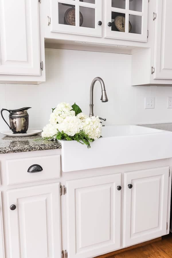 apron front farmhouse sink in a kitchen with fresh white hydrangeas and a brushed nickel faucetwith painted greige cabinets next to a tarnished silver pitcher and vintage scales in glass cabinets above the sink