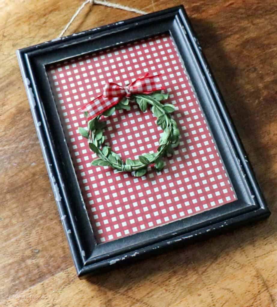 black frame sitting on a wooden table filled with red check wrapping paper with a small christmas wreath glued to the glass
