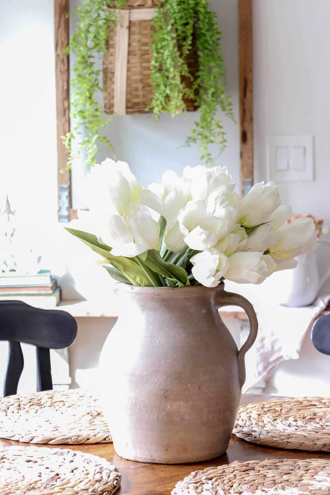 eat in kitchen with woven shades and clear hanging pendant light and a round table with black chairs. A vintage pottery vase is sitting on the table filled with tulips