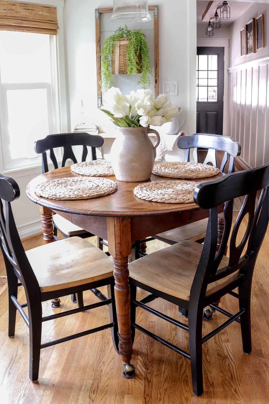 eat in kitchen with woven shades and clear hanging pendant light and a round table with black chairs. A vintage pottery vase is sitting on the table filled with tulips and rattan placemats