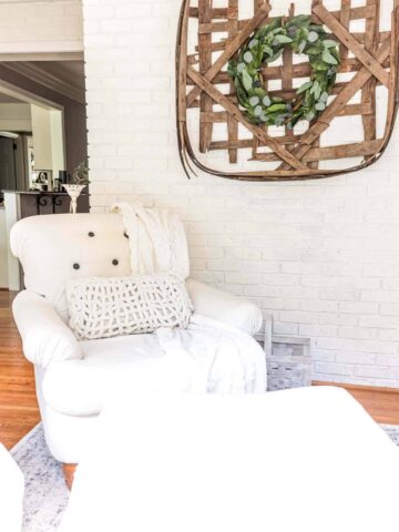 white armchair next to a white lime washed brick wall with a tobacco basket and wreath hanging on the wall