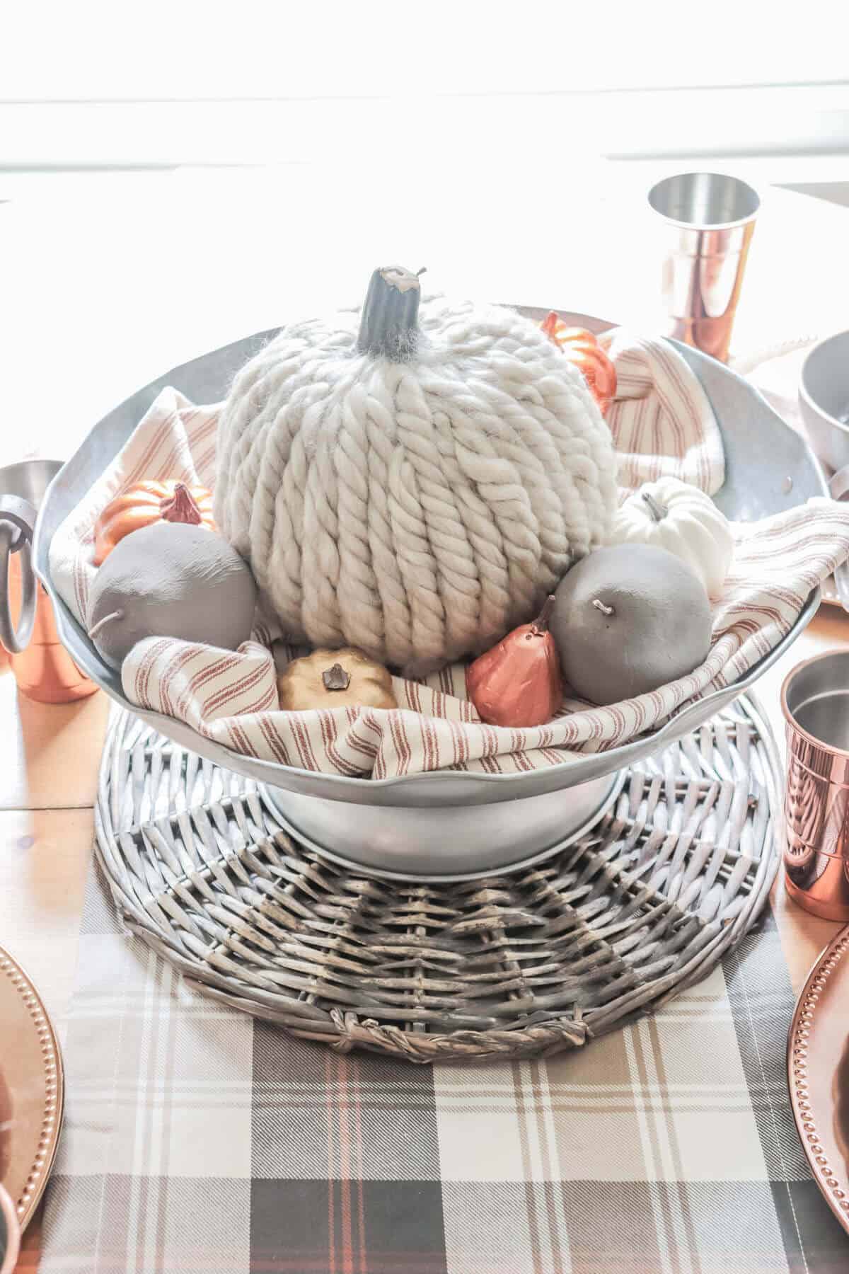 metal centerpiece bowl filled with orange and white striped towel, gray cement pears, a yarn covered pumpkin and copper pumpkins