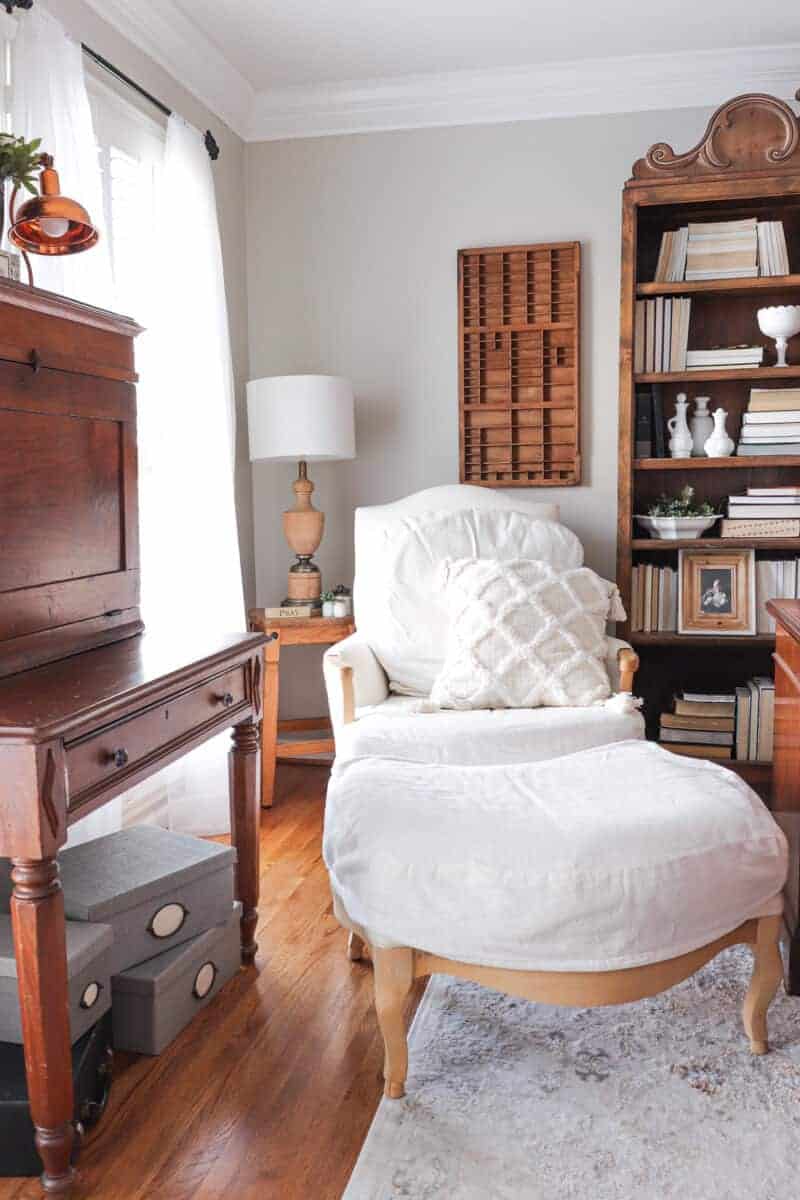 Home office with large white cozy armchair and ottoman next to a bookcase and window.