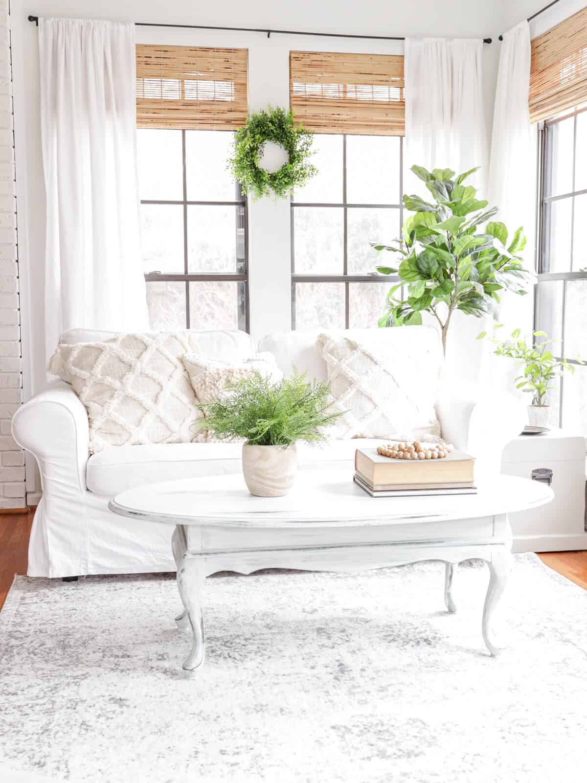 white slipcovered sofa in a sunroom with greenery and woven window shades