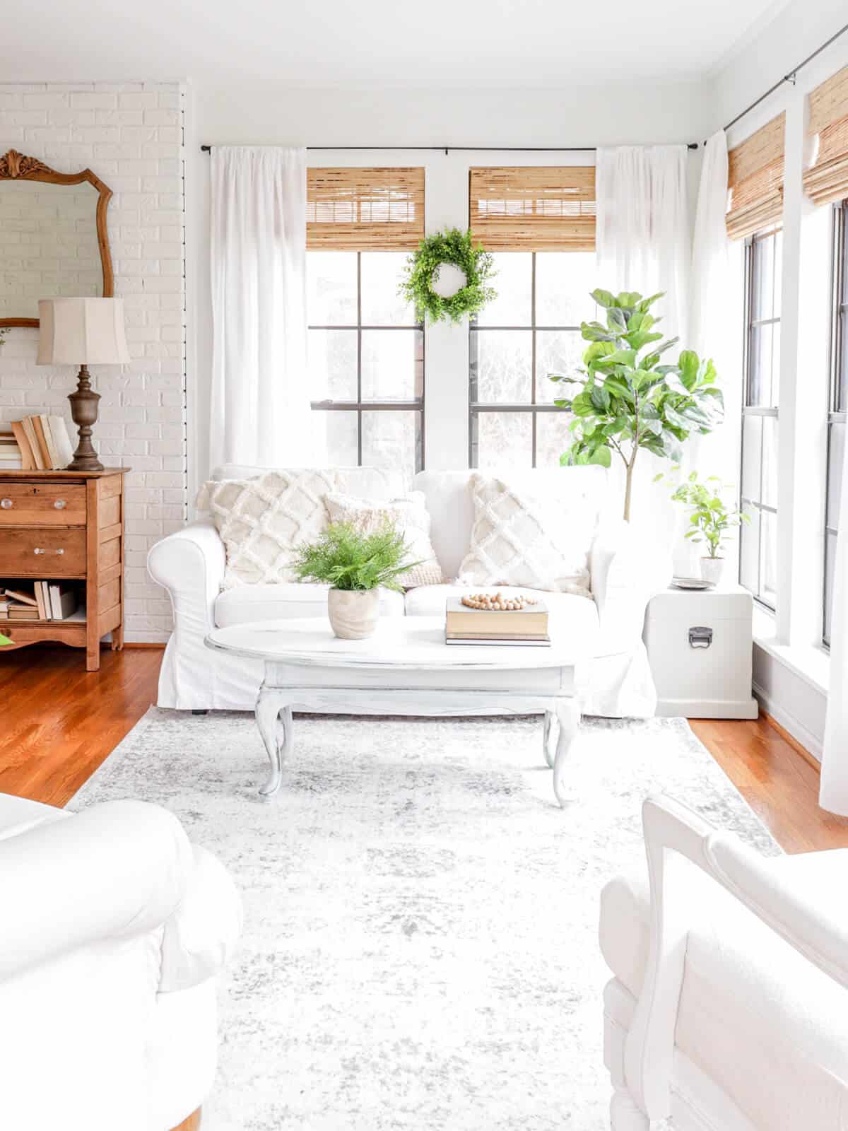 white slipcovered sofa in a sunroom with greenery and woven window shades