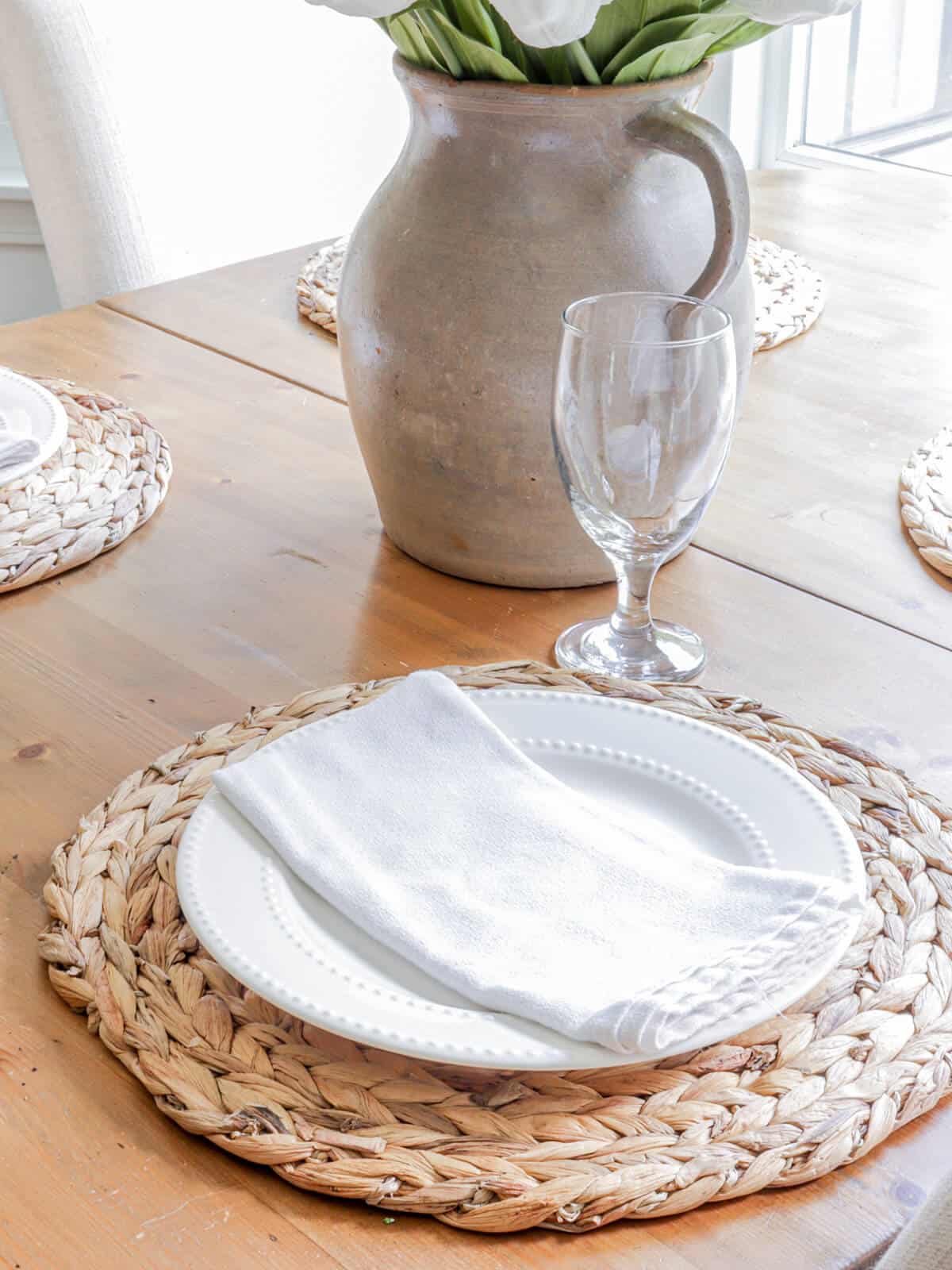simple tablesetting with woven placemats, white plates and napkins and a pottery pitcher filled with faux tulips