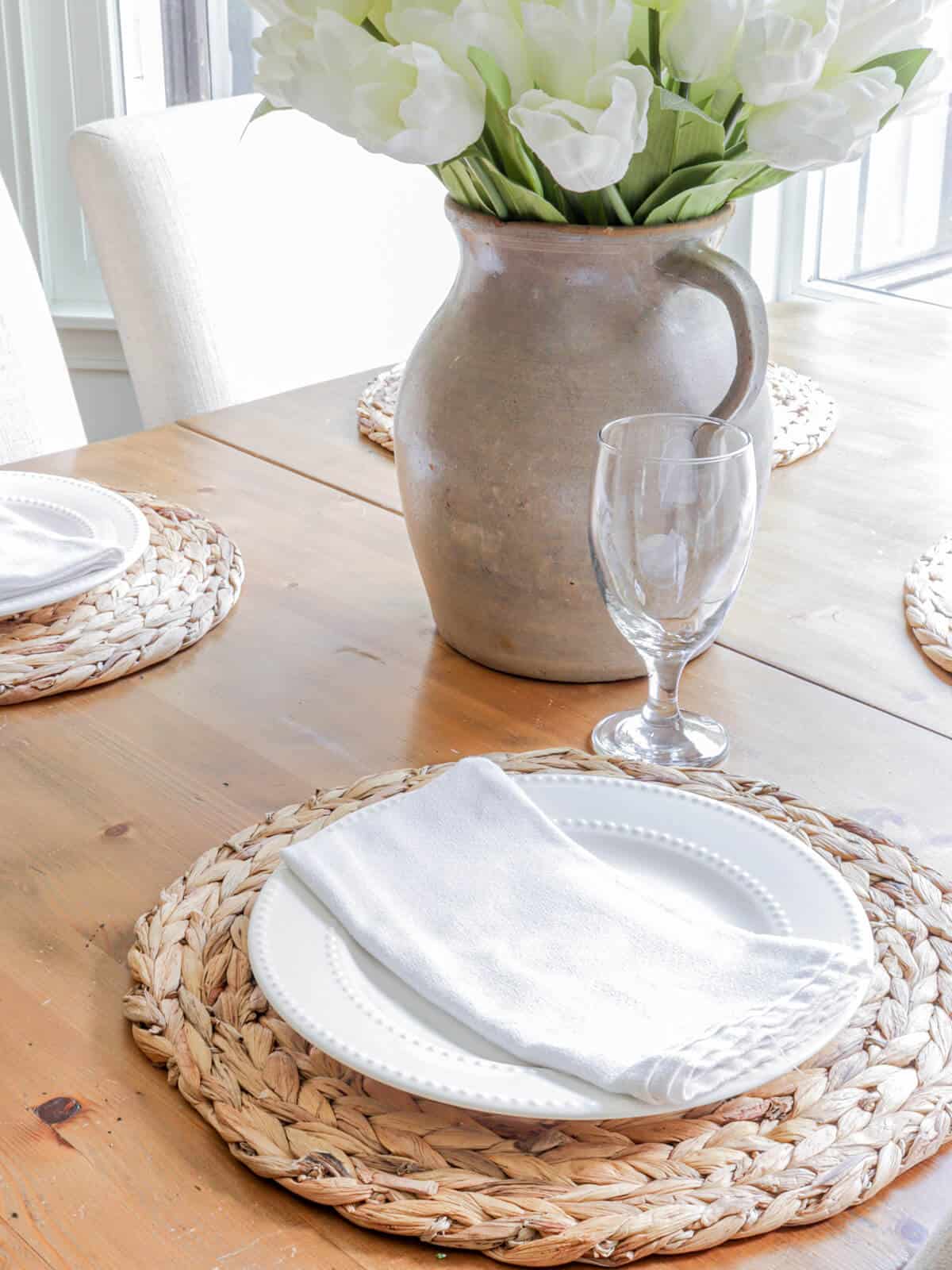 simple summer table setting with woven mats white plates and napkins with a vintage crock filled with tulips