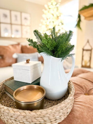 coffee table tray sitting on a tufted ottoman in front of a Christmas tree with a glass vase filled with greenery, a copper candle, books and a woven box