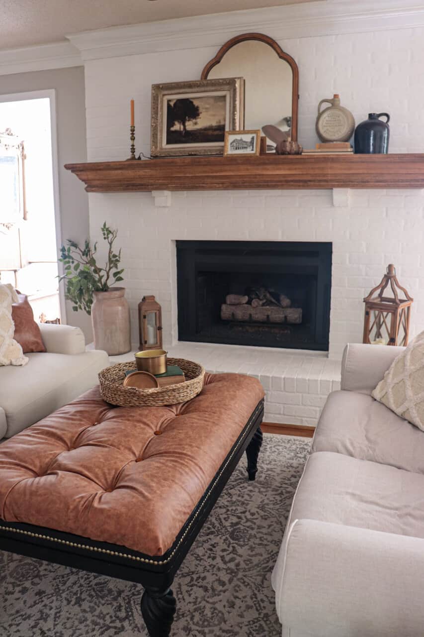 lime slurry fireplace with wood mantel and an old mirror with a framed vintage fall print next to a brass candle with a leather ottoman in front.