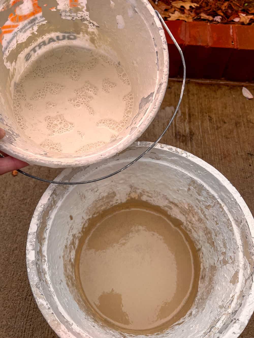 A person holding a bucket of limewash mix in front of a second bucket of a different color.